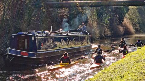 A blue narrowboat is moving along a canal as kayakers pass on the right hand side. There are trees lining the canal.