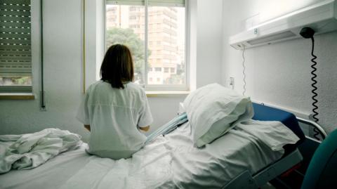 A woman sits in a hospital bed wearing a pale coloured hospital gown. She has her back to the camera and is looking towards a window.
