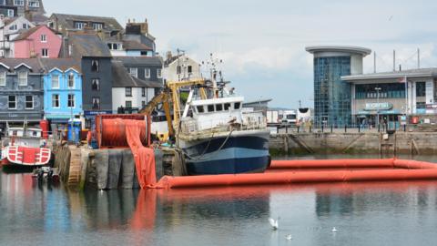 A boat called Accumulate moored up in Brixham Harbour. An orange inflatable boom is around the vessel. The boat is blue and white.