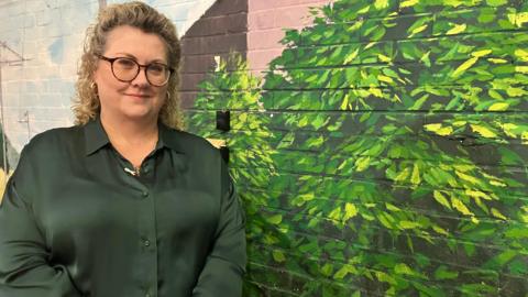 Lisa Squire standing in front of a large mural on a wall. She has long fair hair and is wearing a dark green top. 