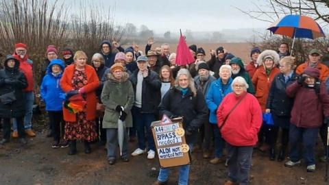 A large group of residents wearing coats and holding umbrellas are gathered by a brown field on a rainy day. One woman at the front of the group is holding a sign that reads: "A33 Chaos. Bypass needed. Aldi not wanted here." 