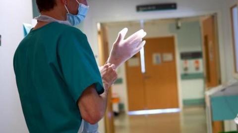 An NHS worker wearing a face mask pulls on a pair of medical gloves while standing in a hospital ward