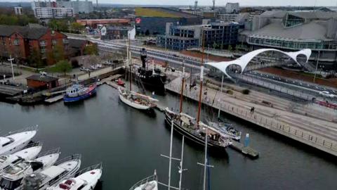 An aerial view shows a marina with white yachts and two tall ships moored at Hull Marina. Modern buildings, a road, and the white Murdoch's Connection bridge line the water’s edge in the background.