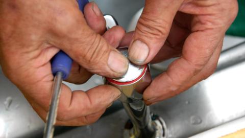 Close up of a plumber's hands on a tap. The plumber is holding two thumbs on the hot tap. A purple screw driver is being held in the right hand. 