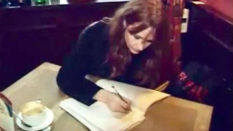 JK Rowling has long brown hair. She is at a wooden table and is writing in a manuscript. She is wearing a black jumper. There is a coffee cup on the table.