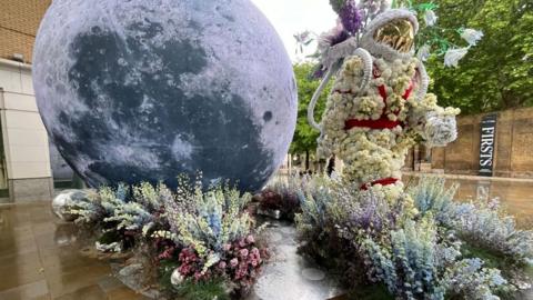A floral installation depicts an astronaut figure facing a large model of the Moon, set among pastel-coloured flowers on a wet pavement in an outdoor urban space.