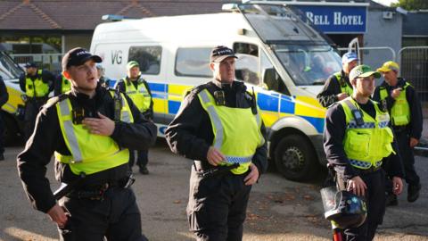A group of police officers in fluorescent tabards speaking into talkback devices and holding riot helmets - there is a large police van in the background at the entrance to a building with a Bell Hotel sign