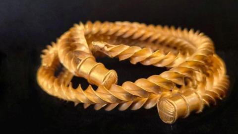 A close up picture of a gold necklace curled around. It looks like a twisted rope with bound ends. 