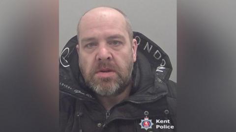 A mugshot of a man looking directly at the camera. He is wearing a black coat. 