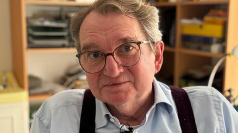 Wearing glasses, a light blue shirt and braces, with short grey hair swept to one side, Jens Hölscher looks at the camera lens as he sits in his study with shelves of books and folders behind him.