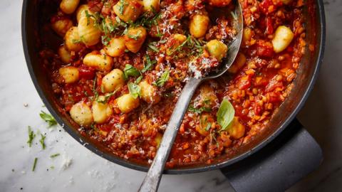 A close up of a cast iron oven pot filled with gnocchi in a tomato lentil sauce with chopped green herbs