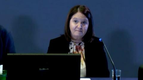 A woman with brown hair wearing a blouse and a black jacket. She is sitting at a desk. 