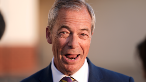 Nigel Farage has short grey hair and his mouth is open as he speaks to reporters, who are not pictured. He is wearing a blue suit jacket and a white shirt.