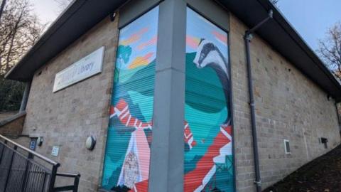 An image of the corner of a square light-coloured stone building, which has shutters at the end of each wall.  The two murals create a single landscape with green hills  and a badger in the foreground and an owl leaning against an upturned book.