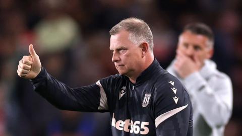 Mark Robins, wearing a Stoke tracksuit, gives a thumbs-up with his right hand, to one of his players 