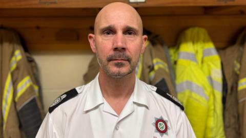 A fire officer wearing a white shirt and black epaulettes is staring at the camera. He has a bald head and short, grey stubble. 