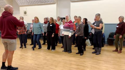 Around 20 women practise as a choir, led by Nick Drew, who wears a burgundy hoodie and beige shorts.