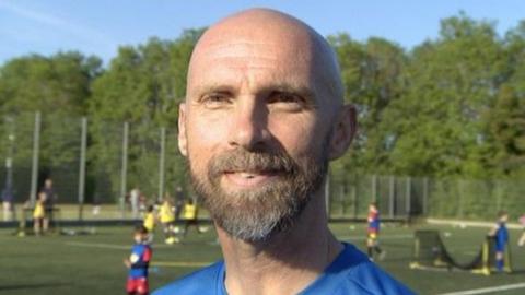 Close-up image of Luke Chadwick. He has a bald head and beard and is smiling at the camera. Behind him are some pitches where children are playing football.