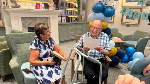 Joe Slyper in a checked shirt smiles as he reads a card, seated beside a smiling woman in a blue and white dress who holds a gift. Behind them are blue and gold balloons and a Happy Birthday banner