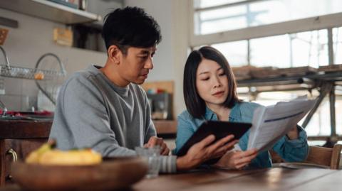 A young couple sit in their kitchen looking at bills. The woman is holding papers and the man is holding a tablet device