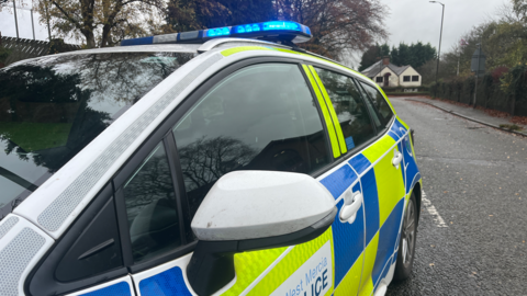 The side of a yellow, blue and white police car with its blue lights on. 