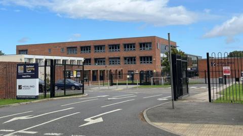 A red-brick, three-storey school building with black windows behind black gates. A grey road runs through the open gates. A sign reads: "Harbour Learning Trust, Healing Academy."