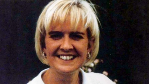 The picture shows a head shot of a smiling woman with a blond bob, wearing a white shirt with earrings 