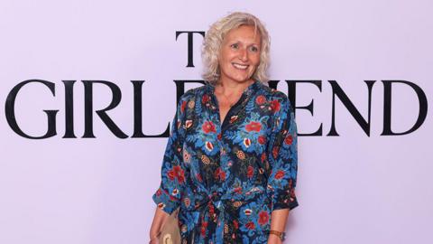 A middle-aged woman in a flowery dress, smiles and poses for a photograph. Behind her is a screen with The Girlfriend written in large letters. 