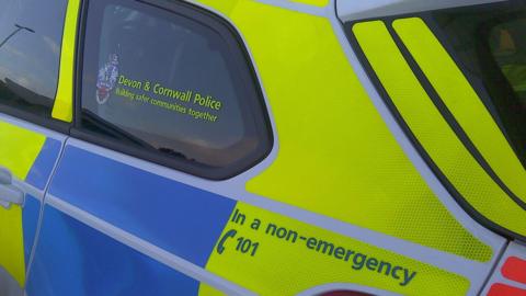 The rear end side of a police car with the Devon and Cornwall Police branding on the side. It has a non-emergency number on the side also. 