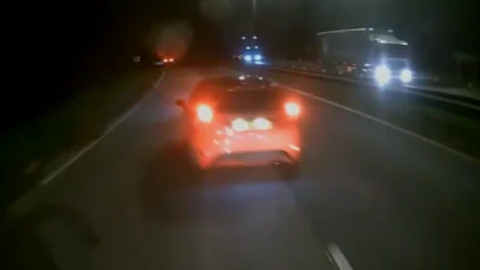 A still picture from footage during a police pursuit involving a red Ford Fiesta on the A11 in Thetford, Norfolk. It is dark and parts of the road are illuminated by headlights. On the opposite side of the road, oncoming traffic includes two lorries which both have their headlights illuminated.
