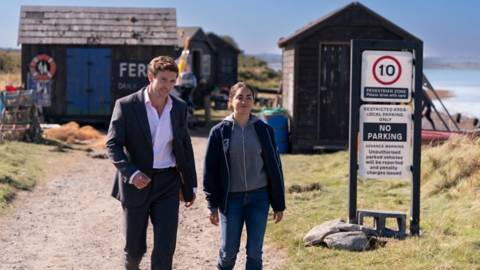 A man and woman walk side by side towards the camera in a publicity shot released by a production company. The man has brown hair and is wearing a suit and white shirt unbuttoned at the neck. The woman is wearing jeans, a grey hoodie and a black jacket. They are on a gravel path, with fisherman's huts behind them.