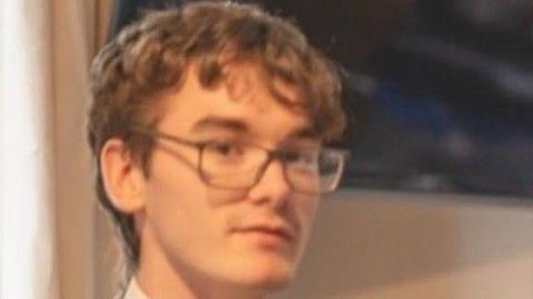 A teenage with short brown hair and glasses looks at the camera