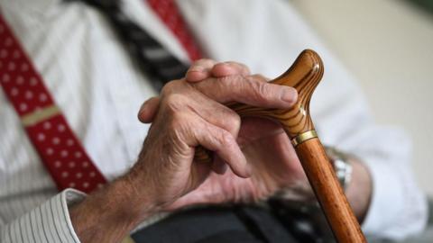 An elderly man sits with a cane in his hand.