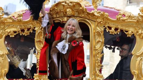 A woman in municipal robes waves from the door of an 18th century golden coach. 