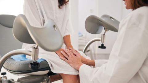 The young woman in a hospital gown sits on the stretcher as the mature doctor in a white lab coat, seated on a stool, holds her hands.