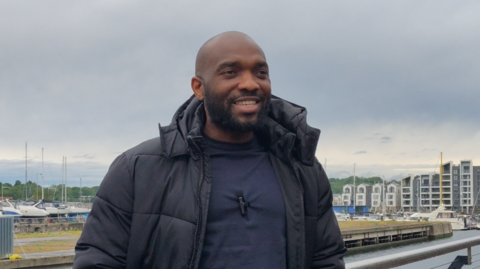 A man stands on a pier. He has a dark blue top and blue jeans on, with a black coat.