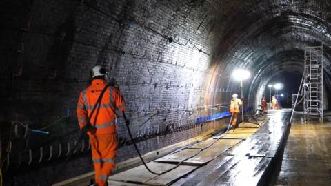 The inside of a tunnel with workers in hi-viz clothing and carrying long cables between them. There is a metal freestanding scaffolding unit and bright spotlights in the otherwise dark tunnel