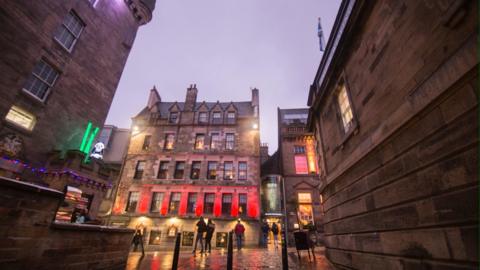 Evening on an Edinburgh street, various coloured lights adorn buildings.