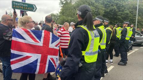 A row of seven police officers stand in a road facing a group of protesters, with trees and a road sign seen behind them.