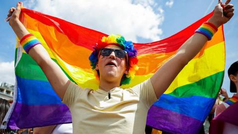 A person standing in the sunshine wearing sunglasses and a rainbow floral headdress, holding a rainbow pride flag up in the air behind their back 