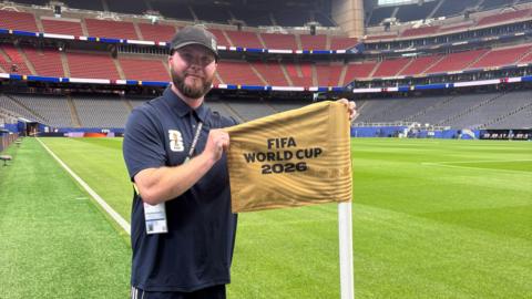 A man in his thirties with a beard and a cap, stood at the corner of a football pitch, holding a yellow flag with FIFA World Cup 2026 printed on it