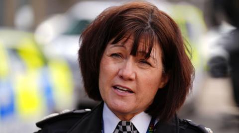 Jo Farrell has short dark brown hair and is mid-speech. She stands in front of a blurred police vehicle and wears a black jacket, white shirt, and black and white checked police tie