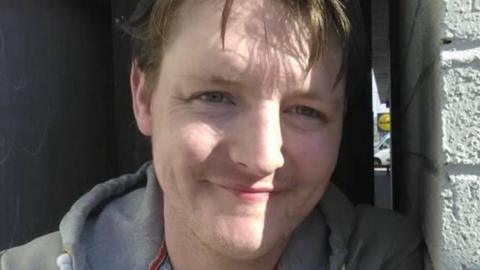 Jamie Forbes leaning against a white wall, smiling at the camera. He is wearing a grey hooded jumper. 