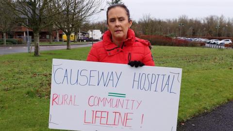 Gemma is wearing a bright red coat and holding a white sign that says Causeway Hospital = rural community lifeline!