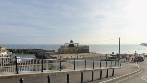 Ventnor beach and harbour wall - it is a calm, sunny day. Cars are parked along the road and people can be seen walking to and from the beach.
