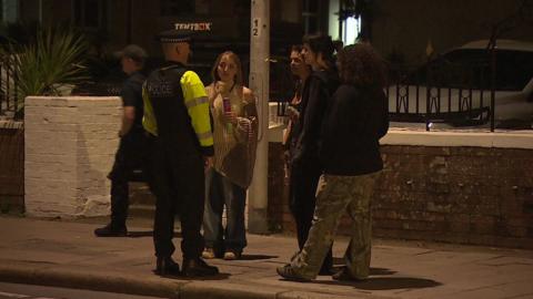 A police officer speaking with three young people standing on a pavement after dark