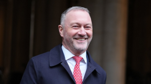 Secretary of State for Housing, Communities and Local Government Steve Reed walks across Downing Street. He is grinning, with closely cropped grey hair and stubble. He wears a navy overcoat, a white shirt and a red paisley tie. 