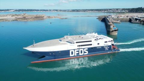 A DFDS ferry coloured white and blue with a red strip at the bottom sailing out of St Helier Harbour. Blue skies and Elizabeth Castle in the background.