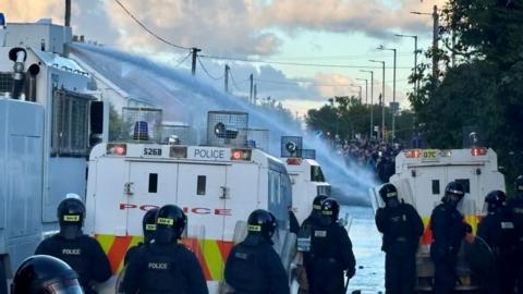 Riot police in the foreground behind police vehicles and a water cannon holding back a crowd in the distance.