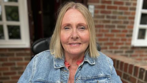 Lynne Warrington has medium length blonde hair and is wearing a denim jacket. She is sat in a black wheelchair and is in front of a residential property.